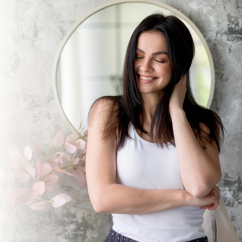 Ellas en Armonía – Bienestar femenino en equilibrio Mujer sonriendo con serenidad frente a un espejo en un espacio luminoso que representa bienestar femenino y equilibrio interior.